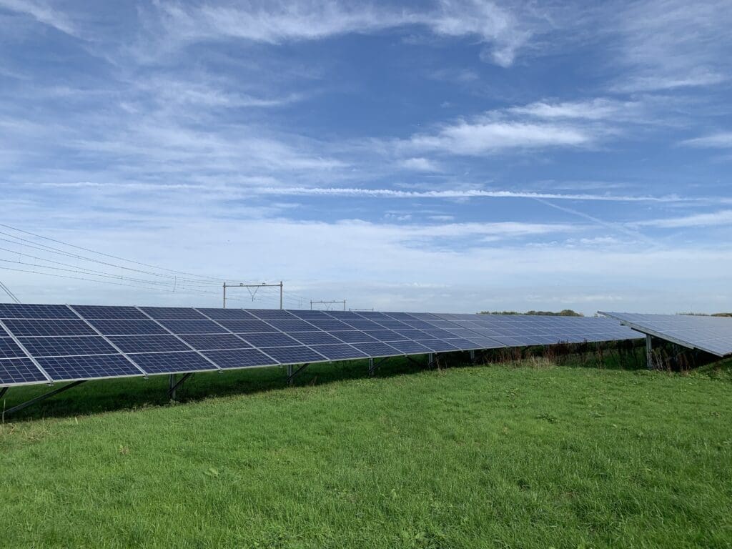 Solar park on a grassland site in the Netherlands, featuring rows of ground-mounted solar panels and high-voltage transmission towers in the background
